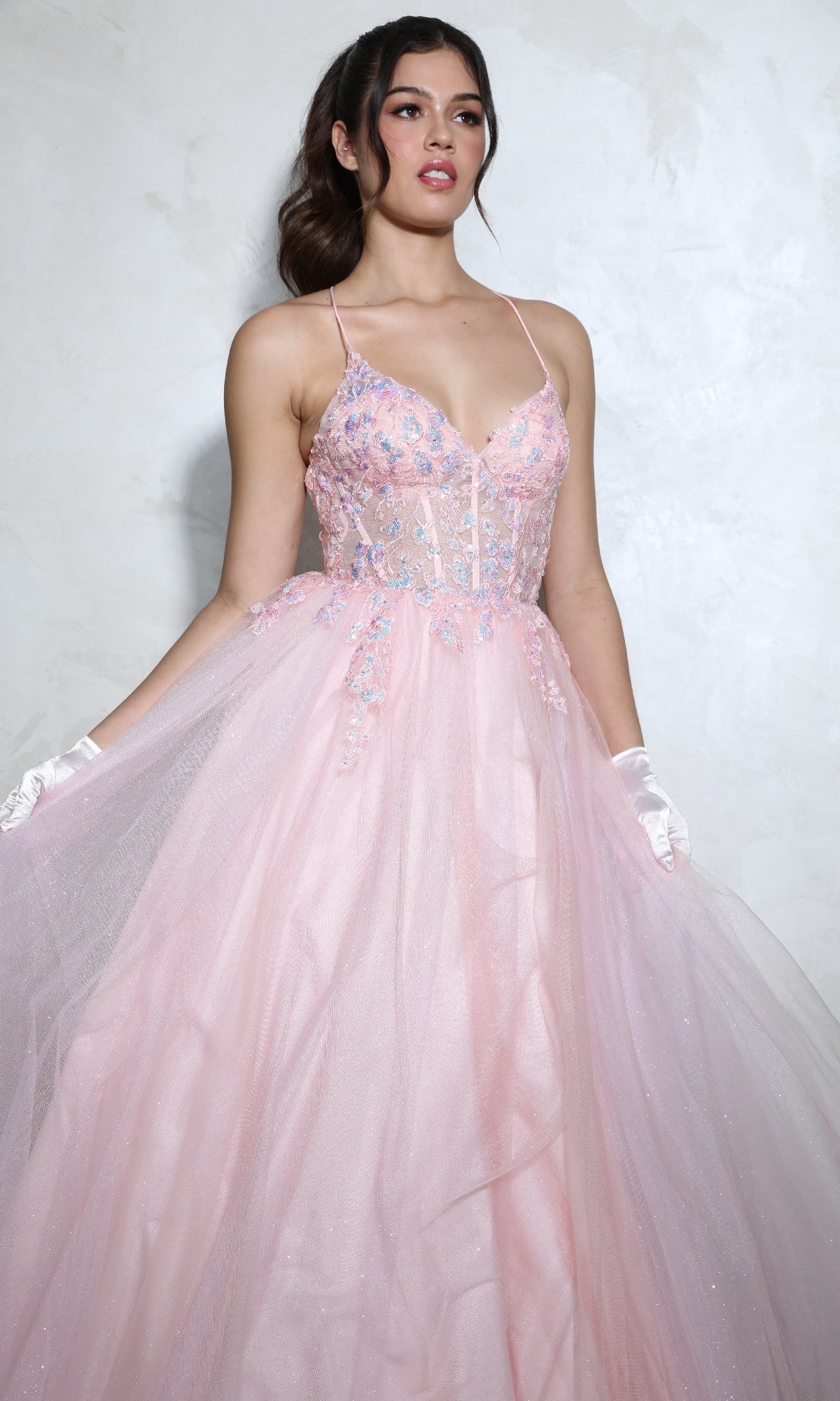 A woman wearing the Eureka 9960 Lace-Up Corset Long Prom Ball Gown, featuring light pink sparkly fabric, floral embroidery, and thin straps, stands against a light textured background.